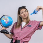 a pleased young woman wearing red shirt in sunglasses holding a globe while flying a blue toy plane on a white background