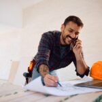 Happy,Construction,Worker,Communicating,On,Cell,Phone,While,Analyzing,Blueprints