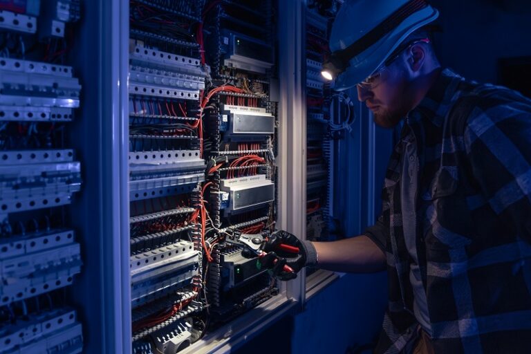 A male electrician works in a switchboard in overalls against the backdrop of emergency lighting.
