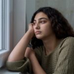 Young woman with curly hair resting her head on her hand while looking out a window with a somber expression.