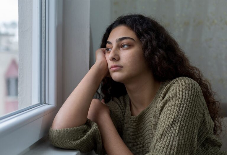 Young woman with curly hair resting her head on her hand while looking out a window with a somber expression.