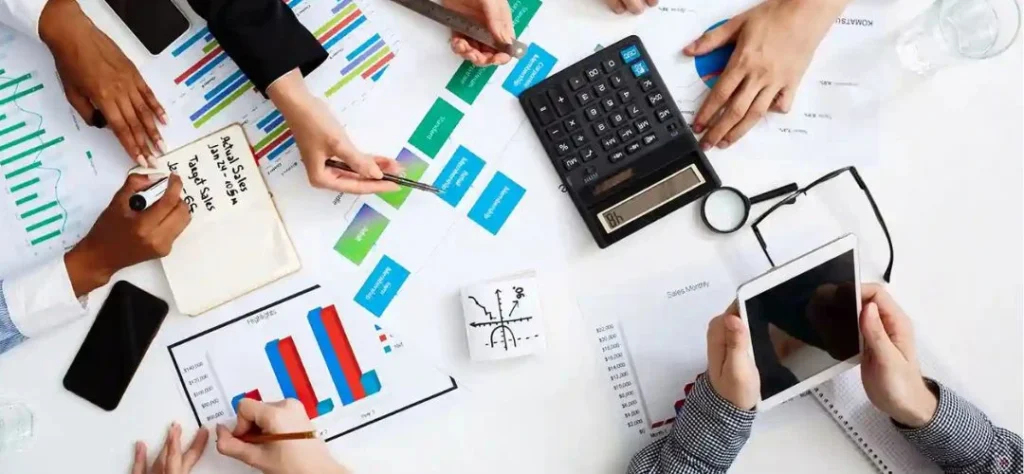 Top-down view of a business meeting with several people reviewing charts and financial documents on a table. Hands are seen using calculators, smartphones, a tablet, and writing notes while discussing colorful graphs and data reports.
