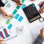 Top-down view of a business meeting with several people reviewing charts and financial documents on a table. Hands are seen using calculators, smartphones, a tablet, and writing notes while discussing colorful graphs and data reports.