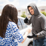 Masked man pointing a gun at a woman during an armed robbery near a car