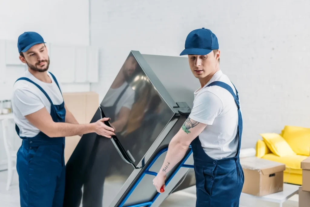 two-movers-in-uniform-using-hand-truck-while-transporting-refrigerator-in-apartment