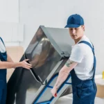 two-movers-in-uniform-using-hand-truck-while-transporting-refrigerator-in-apartment