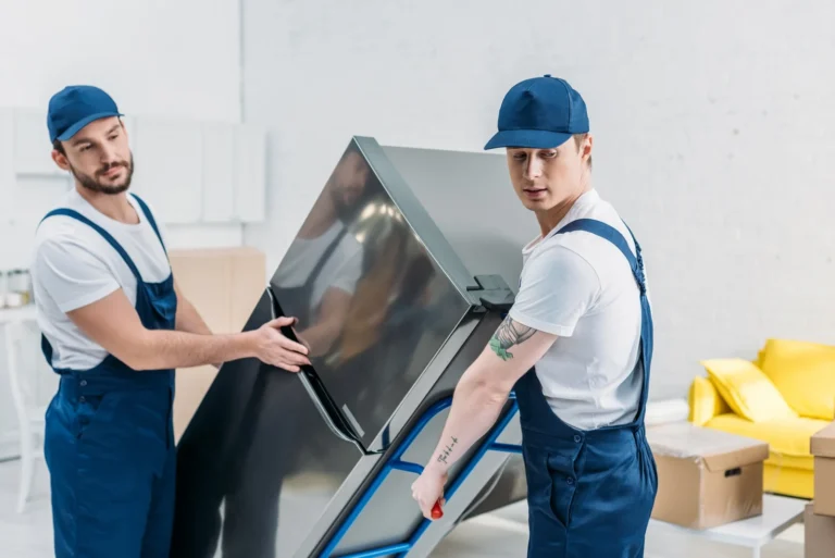 two-movers-in-uniform-using-hand-truck-while-transporting-refrigerator-in-apartment