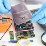 Technician wearing blue gloves repairing a smartphone by inspecting its internal components and circuit board using electronic repair tools.